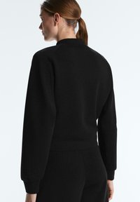 Kvinde med brunt hestehale iført en tætsiddende sort ribstrikket sweater og matchende bukser, set bagfra mod en lys baggrund.