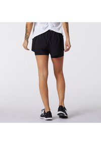 Pantalones cortos atléticos negros con forro interior, cintura elástica y un ajuste relajado. Combinados con zapatillas negras sobre un fondo liso.