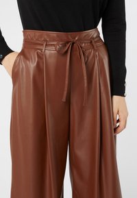 Pantaloni marroni a vita alta in ecopelle con cintura annodata, indossati con un top nero a maniche lunghe, una mano nella tasca e dettagli in bottoni sulle maniche.