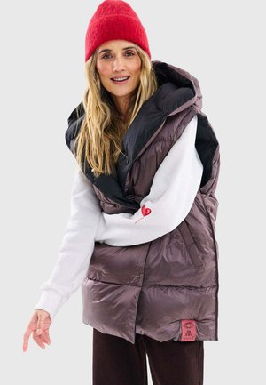 Kobieta ubrana w czerwoną czapkę z daszkiem, białą bluzę z detalem w kształcie serca oraz brązową kamizelkę puchową bez rękawów, stojąca na tle jednolitego tła.