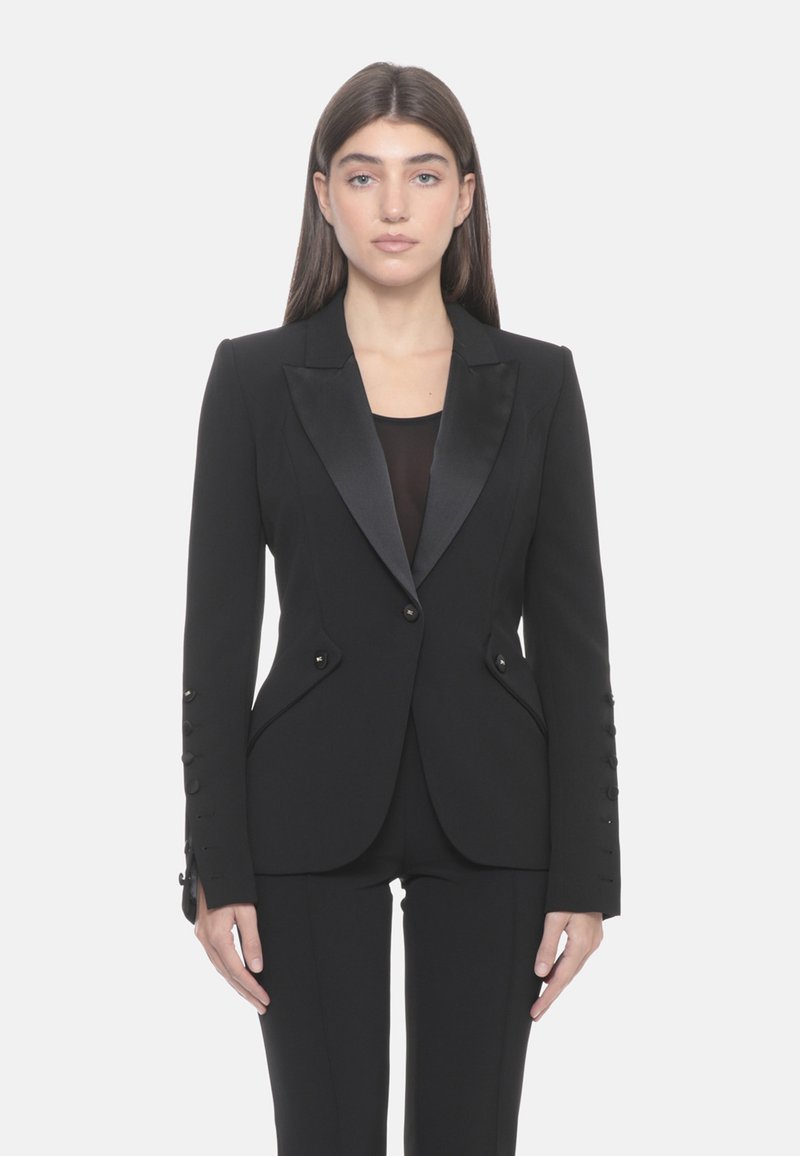 Blazer nero su misura con rever in raso, design aderente e dettagli con bottoni sulle maniche. Abbinato a una maglia nera e pantaloni coordinati.