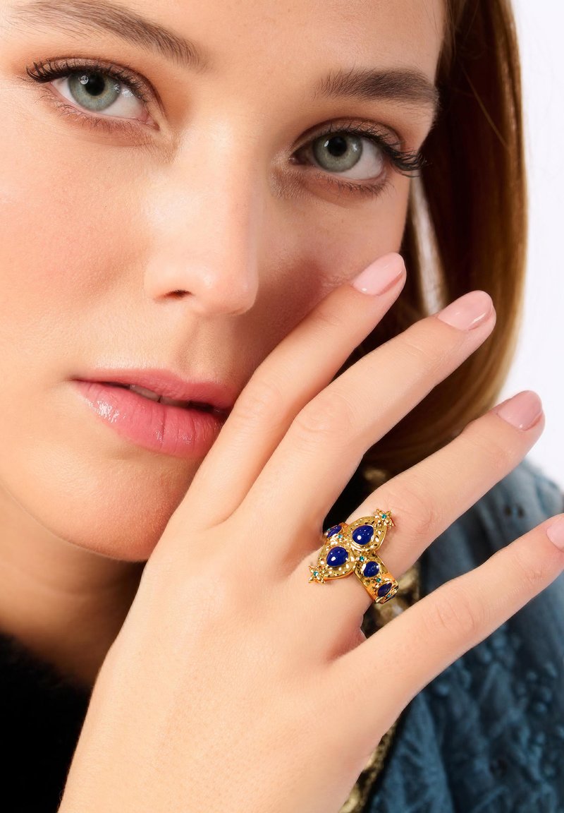 Bague en or ornée de pierres précieuses bleues en forme de croix, avec des détails complexes et de petites touches de vert. La main du modèle est délicatement placée près de son visage.