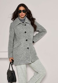 Vrouw in een grijze button-upjas, grijs patroonbroek, zwarte zonnebril, en met een zwarte handtas tegen een lichte achtergrond.