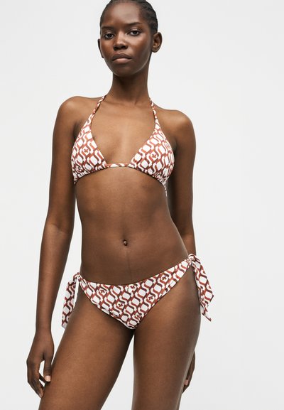 Γυναίκα που φοράει μπικίνι τύπου halter καφέ και λευκό με μοτίβο, με κάτω μέρος που δένει στο πλάι, στέκεται μπροστά σε απλό ανοιχτό φόντο.