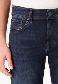 Tamno plavi traperice s uskim krojem, s kontrastnim šavovima, dugmetima na prednjem dijelu i prednjim džepovima. Glatka tekstura, minimalistički dizajn.