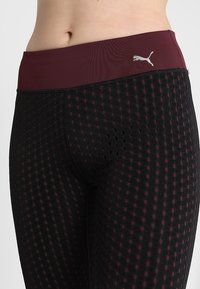 Zbliżenie na talię i biodra w czarnych legginsach z bordowym pasem i perforowanym wzorem w kropki, z logo Puma w kolorze srebrnym na pasie.