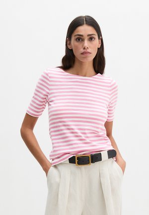 Femme aux cheveux raides et foncés portant un haut à manches courtes rayé rose et blanc, un pantalon plissé blanc et une ceinture noire avec une boucle dorée.