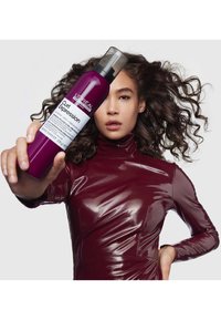 Produkt stylizujący Curl Expression od L'Oréal w fioletowej butelce z pompką, trzymany przez modelkę w błyszczącej burgundowej bluzce.