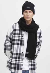 Zwarte beanie en een lange zwarte sjaal gecombineerd met een zwart-wit geruite shirt. Het materiaal lijkt zacht; het shirt heeft een knoopsluiting en logo-detail.