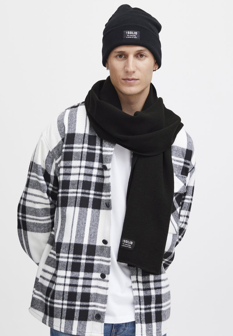 Zwarte beanie en een lange zwarte sjaal gecombineerd met een zwart-wit geruite shirt. Het materiaal lijkt zacht; het shirt heeft een knoopsluiting en logo-detail.