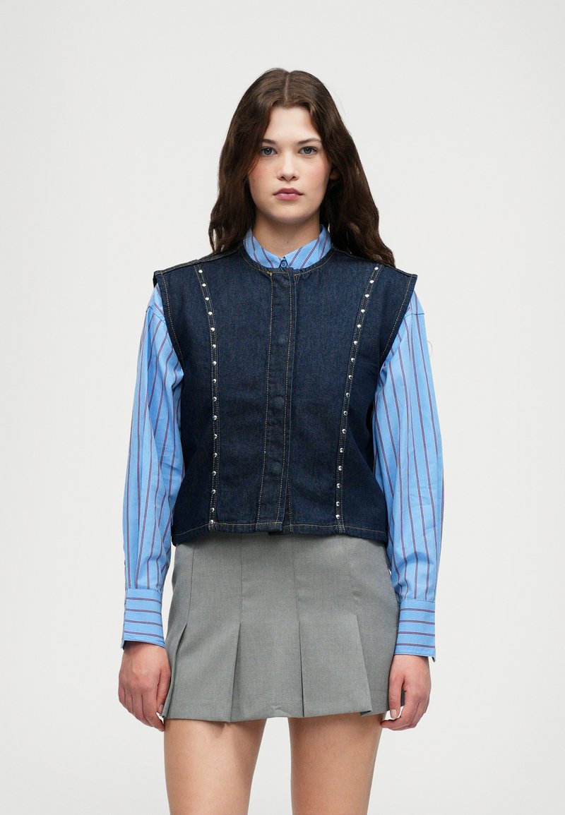Džínová vestu s kovovými hrotmi, skrátený strih, tmavomodrá farba. Nosená cez modro-biele pruhované tričko s dlhým rukávom a šedú plisovanú sukňu.