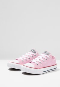 Różowe płócienne trampki z białymi sznurowadłami i gumowymi podeszwami. Posiadają teksturowaną powierzchnię, białą osłonę palców oraz cienką czerwoną i granatową paski.