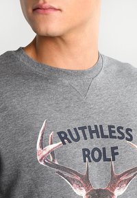 Felpa grigia con scollo rotondo. Presenta il testo "Ruthless Rolf" e un design di corna illustrate. Tessuto con una texture liscia, vestibilità comoda.