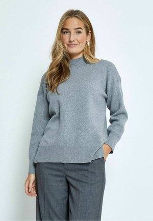 Grey knitted sweater with a high neck and ribbed hem, featuring a relaxed fit and dropped shoulders, paired with grey trousers.