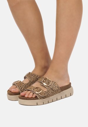 Piedi con sandali slip-on color beige con motivo maculato nero e due fibbie dorate su suole spesse beige.