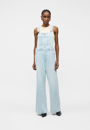 Ženska, oblečena v svetlo modre jeansa kombinezon in brezrokavnik bež barve, stoji ob enobarvni svetlo sivi ozadju.