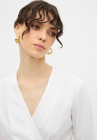 Blusa bianca avvolgente con scollatura a V profonda e maniche lunghe, realizzata in tessuto morbido. Il modello indossa grandi orecchini a cerchio dorati.