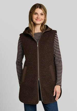 Woman wearing a brown textured zip-up sleeveless vest over a brown and white striped long-sleeve shirt against a gray background.
