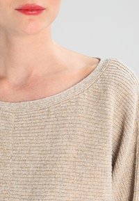 Parte inferior de la cara y el cuello de una mujer que lleva un suéter de punto texturizado beige con un escote ancho, contra un fondo gris claro.