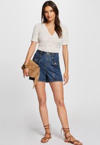 Weißes Strickoberteil mit V-Ausschnitt, hoch taillierte Jeansshorts in Dunkelblau und eine beigefarbene Wildleder-Crossbody-Tasche mit Fransenakzenten. Braune Riemchensandalen.