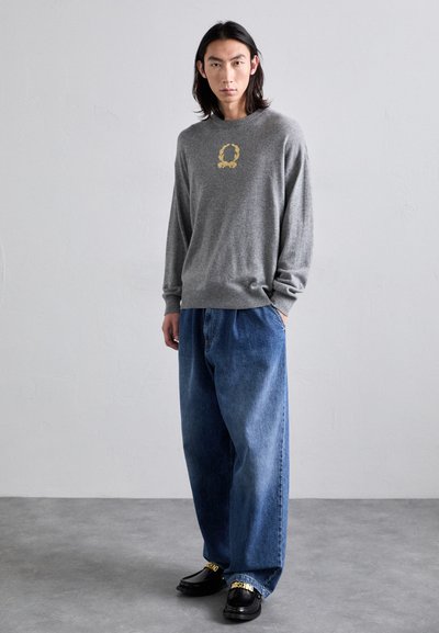 Grau gestrickter Pullover mit einem goldenen Logo, kombiniert mit lockeren blauen Denim-Jeans. Schwarze Schuhe mit goldenen Akzenten runden das Outfit ab.