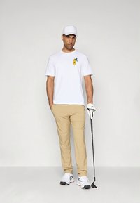 T-shirt branca de algodão com estampa de limão, calças bege, boné branco e taco de golfe. Os sapatos são pretos e brancos com detalhes em vermelho.