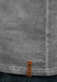Graues Baumwoll-T-Shirt mit einer weichen Textur, featuring einem genähten Saum und einem braunen Etikett, das unten links "pilots!" zeigt.