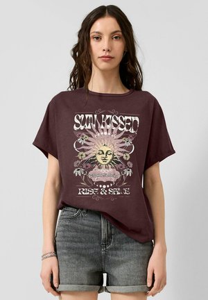 Jonge vrouw die een paars T-shirt draagt met een zonafbeelding en de woorden "Sun Kissed", gecombineerd met opgerolde grijze spijkershort.