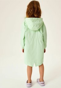 Hellegrüner Regenmantel mit Kapuze, tailliert und mit langen Ärmeln. Die Rückansicht zeigt ein knielanges Design mit gespaltem Saum. Kombiniert mit lavendelfarbenen Clogs.