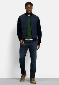 Marineblaue gesteppte Jacke mit gerippten Ärmeln über einem grünen Pullover, kombiniert mit dunklen Jeans und hellbraunen Wanderschuhen. Frontalansicht, minimalistischer Hintergrund.