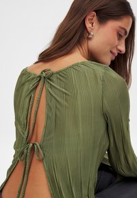 Groene geplooide blouse met een strikdetail aan de achterkant, voorzien van een ronde halslijn en een open rugontwerp. Gladde, zachte stof met een gedrapeerd uiterlijk.