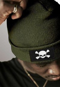 Olive green beanie hat with ribbed texture featuring a black patch with white skull and crossbones design.