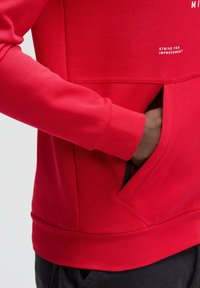Red sweatshirt featuring a textured finish, kangaroo pocket with black lining, ribbed cuffs, and hem. White text reads "STRIVE FOR IMPROVEMENT."