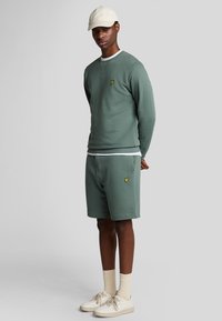 Felpa verde in cotone con maniche lunghe e colletto tondo; pantaloni corti abbinati, entrambi con un piccolo logo giallo. Un cappellino chiaro e delle scarpe da ginnastica completano il look.