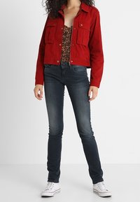 Rode cropped denim jacket met twee voorkruisen, gedragen over een top met luipaardprint, gecombineerd met donkere skinny jeans en witte sneakers.