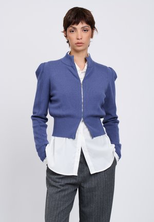 Donna che indossa un maglione blu con zip sopra una camicia bianca e pantaloni grigi a righe sottili, in piedi con le mani in tasca davanti a uno sfondo semplice.