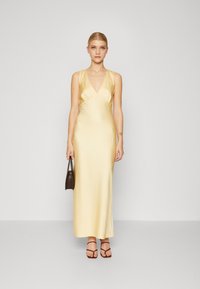 Femeie îmbrăcată cu o rochie maxi din satin galben pal, fără mâneci, ținând în mână o geantă mică de culoare maro închis și purtând sandale cu barete de culoare maro.