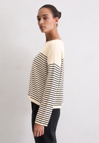 Langarmshirt met witte en marineblauwe strepen en een brede halslijn. Heeft een comfortabele pasvorm en een geribbelde zoom. Model draagt zwarte broek.