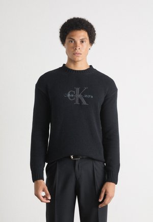Čierny pletený sveter s rebrovanými manžetami a výstrihom. Na prednej strane má logo "Calvin Klein Jeans" vo svetlo sivej farbe. V kombinácii s čiernymi nohavicami.