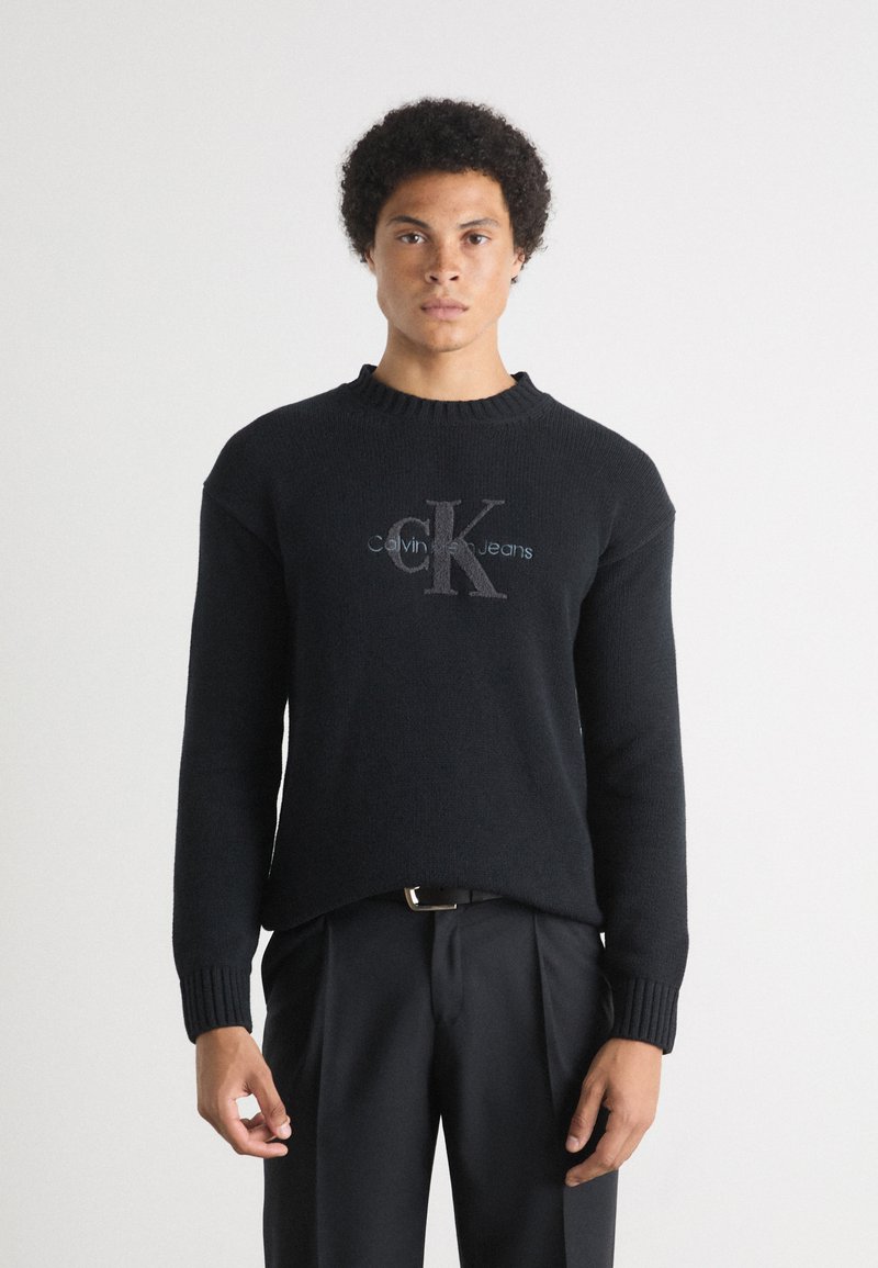 Juodas megztinis su rausvomis apykaklėmis ir rankogaliais. Pavyzdžiui, priekyje yra šviesiai pilkas "Calvin Klein Jeans" logotipas. Suderinta su juodomis kelnėmis.