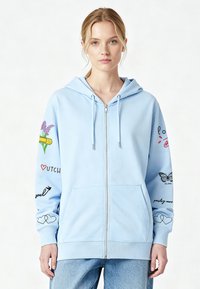 Sweat à capuche bleu clair avec fermeture éclair, fabriqué en tissu doux. Présente des motifs colorés imprimés sur les manches, incluant des fleurs, des papillons et des accents de texte.
