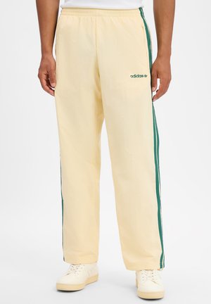 Beige Adidas-Trainingshose mit grünen Seitenstreifen, elastischem Bund und Logo am linken Oberschenkel, getragen mit weißen Sneakers.