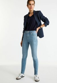 Jeans ajustados de color azul claro con cintura alta, combinados con una blusa azul marino de cuello en V y un blazer oscuro. El calzado consiste en zapatillas blancas con detalles en negro.