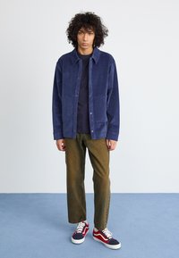 Navy corduroy shirt jacket with front pockets, worn over a dark top. Olive green trousers and red, navy, and white sneakers complete the look.