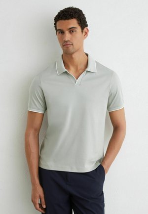 Man die een lichtgrijs poloshirt met korte mouwen en een donkere broek draagt, staand tegen een effen witte muur met één hand in de zak.