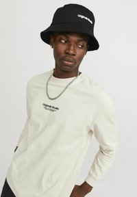 Long-sleeve beige shirt with "Originals Studio" logo, paired with a black bucket hat featuring white text and a silver chain necklace.