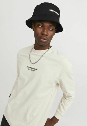 Beige lange mouwen shirt met het "Originals Studio" logo, gecombineerd met een zwarte bucket hat met witte tekst en een zilveren ketting halsketting.