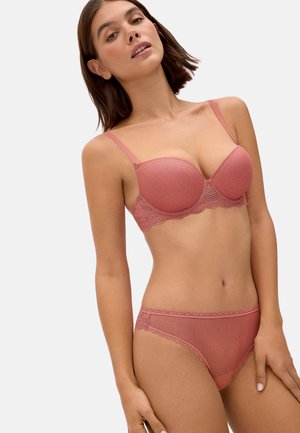 Mujer con conjunto de lencería de color rosa con detalles de encaje, posando contra un fondo liso y claro.