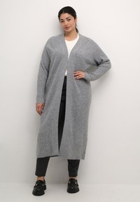 Cardigan lungo grigio a coste con apertura frontale, lavorazione a maglia texturizzata, vestibilità rilassata e maniche lunghe, indossato sopra una maglietta bianca e pantaloni neri.