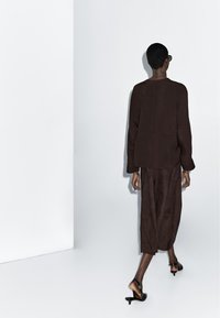 Brązowy oversize'owy top i pasująca długa spódnica; gładka faktura. Czarne sandały na obcasie z paskiem i klamrą. Widok od tyłu.